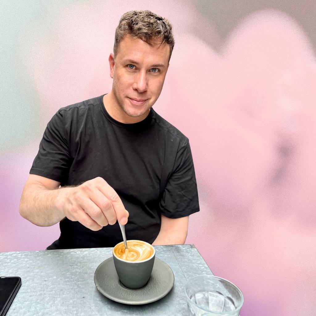 Nicholas James, Founder of Blooming Checkout in a black t-shirt stirring a cup of coffee with a blurred background